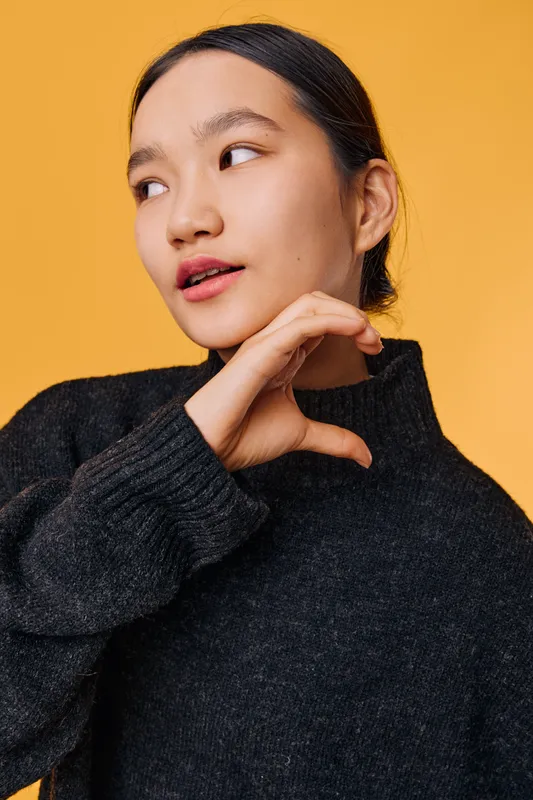 Young Asian woman in black sweater posing on orange background with hand near face, looking sideways, natural makeup and casual hairstyle for beauty or fashion portrait.