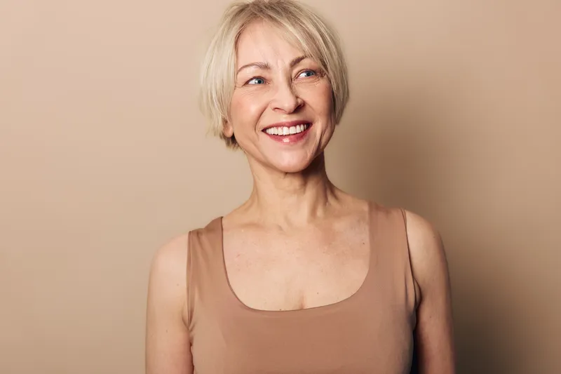 Smiling middle aged woman with short blonde hair wearing sleeveless top looks away joyfully against beige background. Concept of happiness, confidence, natural beauty, and positive mood.