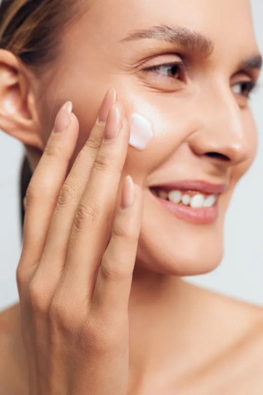 A close up of a smiling woman applying cream to her cheek with fingers. The image focuses on skincare, beauty routine, and healthy skin care concepts.