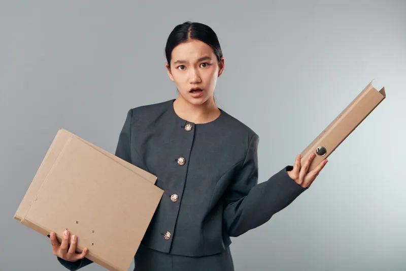 Woman holding cardboard boxes in each hand with confused expression on gray background. Businesswoman in professional suit stressed with shipping parcels for delivery or storage task.