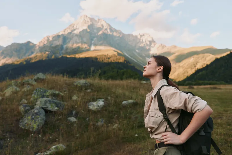 Young woman with a backpack enjoys fresh mountain air while standing in a grassy field with rocky terrain and a towering peak in the background under a partly cloudy sky.