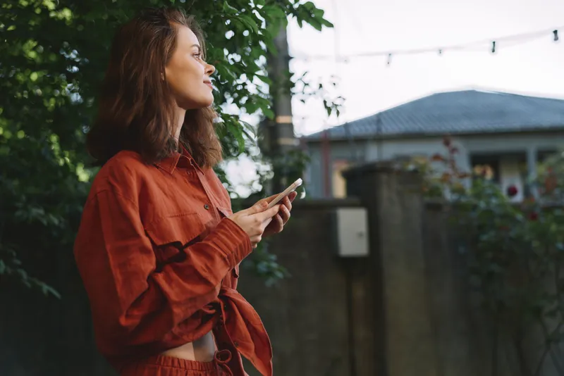 Woman in a red shirt outdoors, looking away while holding a smartphone. Natural daylight, garden backdrop, lifestyle and casual moment. Enjoying a calm, sunny vibe