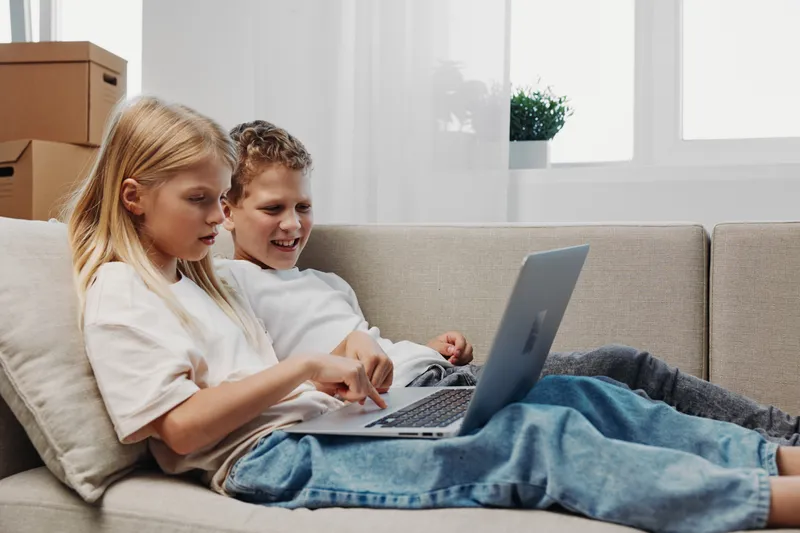 Two happy children sitting on sofa using laptop together in cozy living room with natural light from window behind.