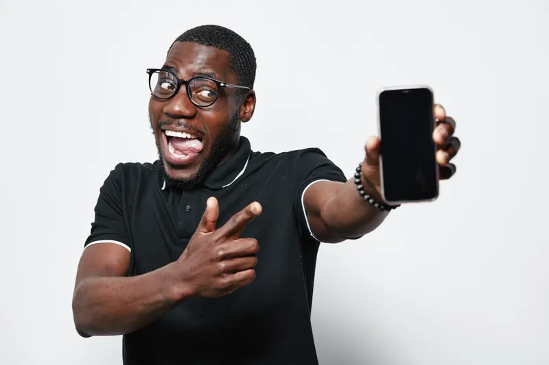 African American man with dark skin and glasses smiling and pointing at smartphone in hand. Casual black shirt, happy expression, isolated on white background for technology concept.