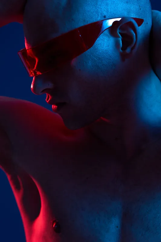 Stylish man posing dramatically with red tinted glasses, showcasing a bold aesthetic in a striking blue and red light setup, emphasizing modern fashion trends.