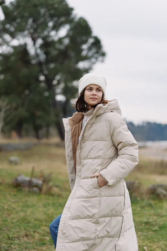 Woman in a beige long puffer coat stands in a grassy field near a lake, winter fashion, warm outerwear, outdoor portrait with natural landscape and calm water.