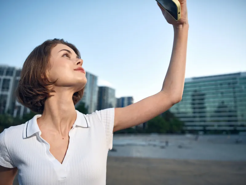 A cheerful female professional takes a selfie outdoors in a modern city setting. She smiles brightly, exuding confidence and energy while capturing a dynamic moment.