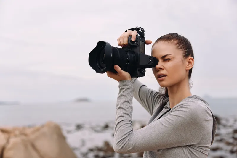Young woman photographing seascape, wearing casual grey hoodie, focused expression, soft natural lighting, outdoor setting, capturing memories near the beach