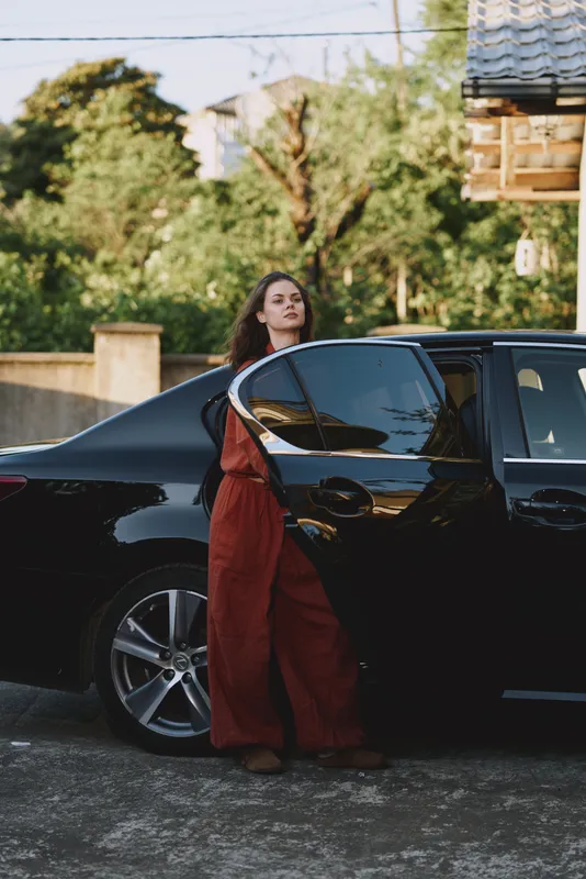 Young woman with brunette hair wearing a red jumpsuit leans against a sleek black car on a city street in warm late afternoon light, posing with confidence and street style