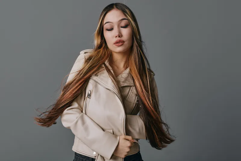 Young woman with long hair and closed eyes wearing beige jacket posing indoors on gray background for fashion portrait or casual style photography with natural expression.