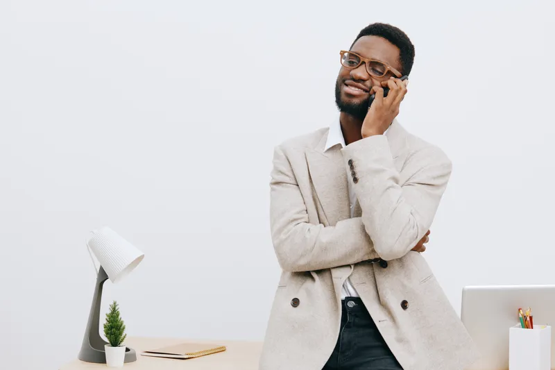 Professional young african-american man in beige blazer and glasses talking on mobile phone, standing in modern office with white background, laptop and desk accessories, casual business look, communi