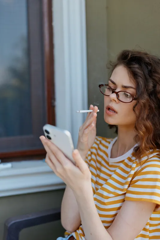 Woman in casual striped shirt outdoors, focused on smartphone, cigarette in hand, relaxed mood, natural light, candid lifestyle moment, everyday scene and calm atmosphere