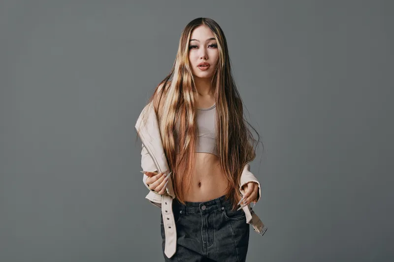 Young woman with long hair wearing crop top and jacket posing with hands in pockets standing against plain gray background in studio.