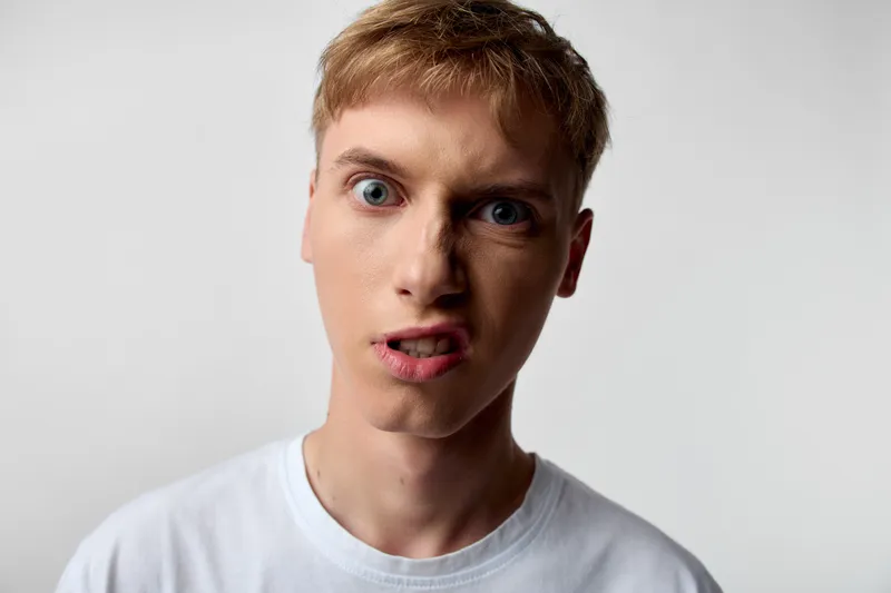 Closeup of a surprised man with wide eyes and a puzzled expression, wearing a whitetshirt, in a studio with a neutral backdrop, conveying confusion and curiosity for stock listing