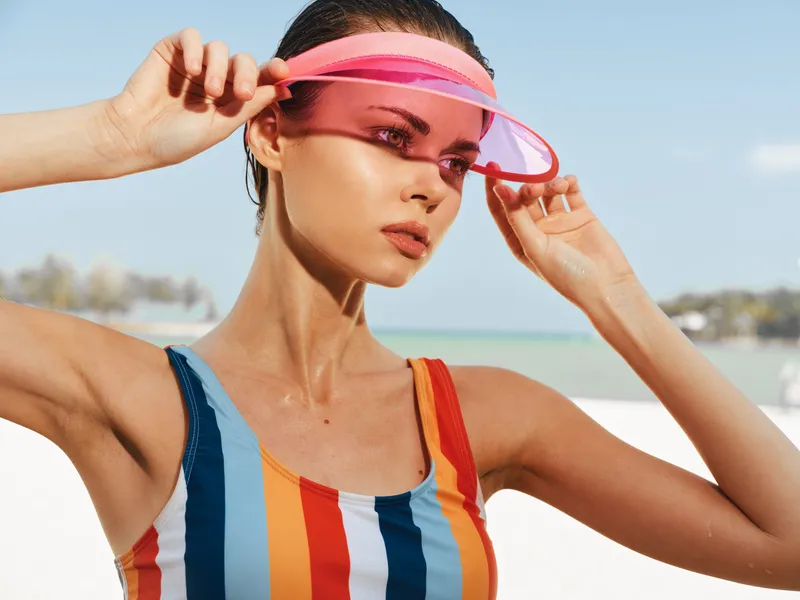 Portrait of a young woman at the beach wearing a striped swimsuit and pink visor, with sunlit skin and hair tied back, conveying bold summer style and confidence.