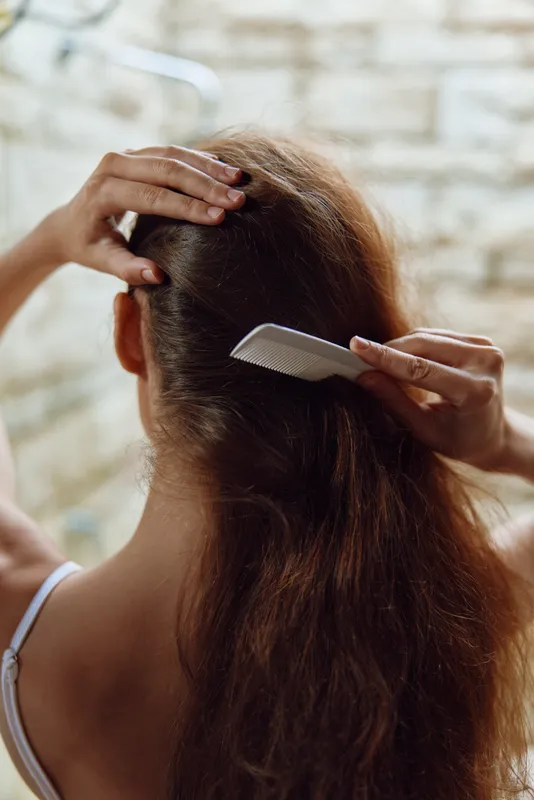 woman combing hair with white plastic comb indoors near tiled wall, long brown hair, personal care and grooming, natural light, casual setting