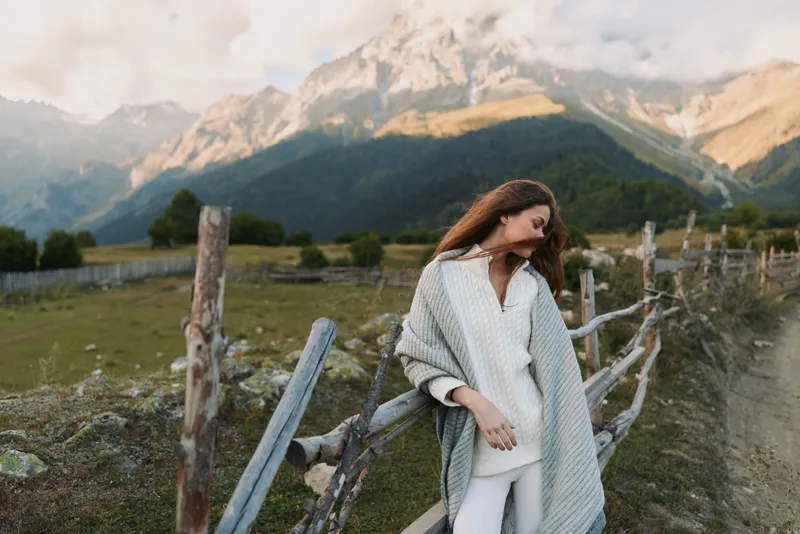 Young woman wrapped in a knitted blanket leans on a wooden fence in a mountainous rural landscape during a calm autumn day.