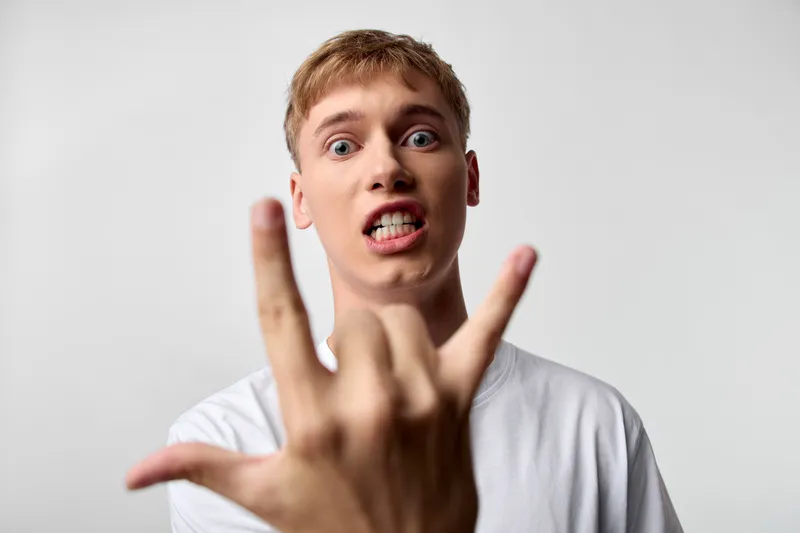 Front view of a young man making a bold hand gesture with an expressive face, wearing a white t shirt in a studio shot, creating an edgy, rebellious vibe and strong nonverbal message.