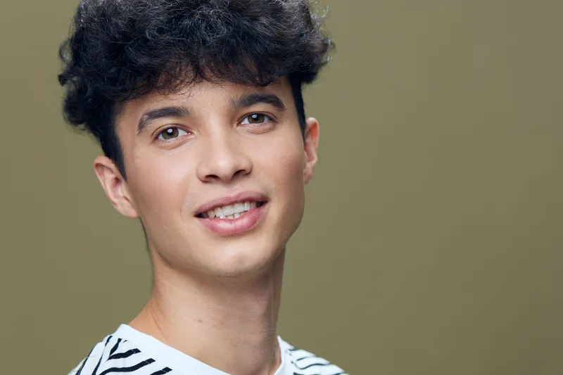 smiling young man with curly hair wearing striped shirt, looking at camera against olive green background in bright soft light