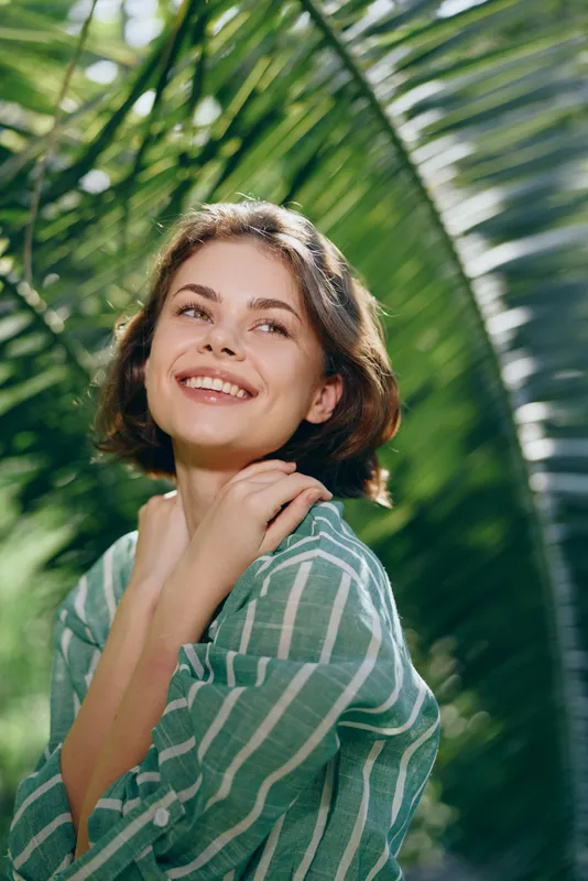 Portrait of a smiling woman outdoors, bathed in soft sunlight, with lush palm leaves in the background and a striped shirt.