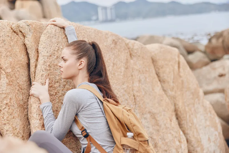 Young female climber with brown hair in a ponytail, wearing gray athletic wear and a brown backpack, climbing rocky terrain near the coast