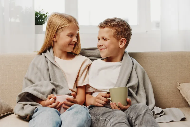 Two happy children wrapped in cozy blankets sit on a sofa holding mugs, smiling at each other in a bright living room. Concept of warmth, comfort, friendship, and family bonding.