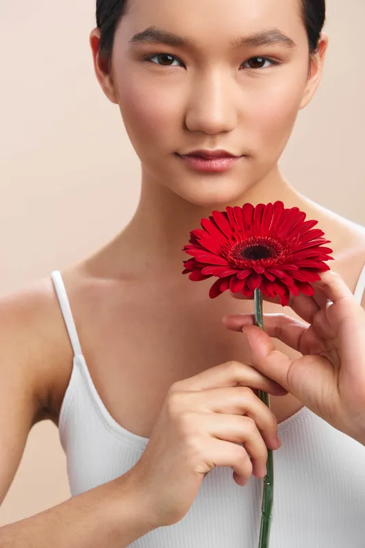 asian woman gazes softly while holding a red flower against a neutral backdrop, exuding calm elegance and focus, a poised and serene portrait with gentle lighting.