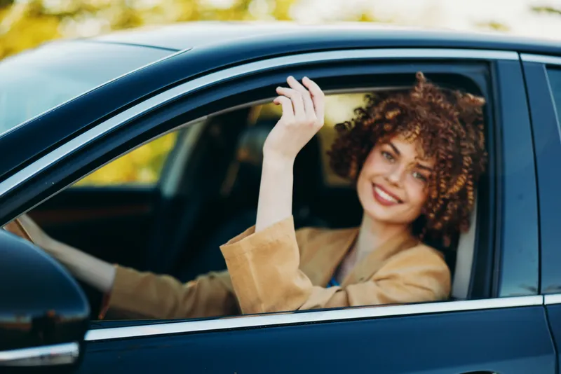 Cheerful woman with curly hair sits in car window, smiles at camera, wearing a beige jacket, casual lifestyle moment captured with warm retro film color and urban street background