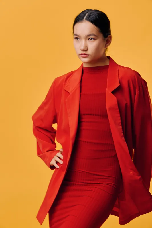 Young woman in red dress and jacket posing confidently with hand on hip against yellow background, fashion portrait with professional and stylish appearance.