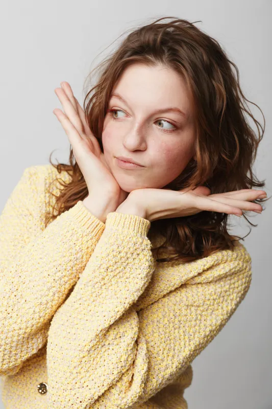 Young woman with wavy brown hair wearing yellow sweater looks sideways with playful expression, hands framing face. Studio portrait on white background, concept of youth and casual style.