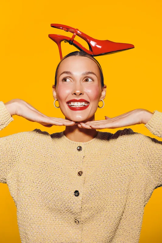 Smiling woman with red high heel shoes on her head poses with hands under chin against bright yellow background. Concept of fashion, fun, and playful mood in studio portrait.