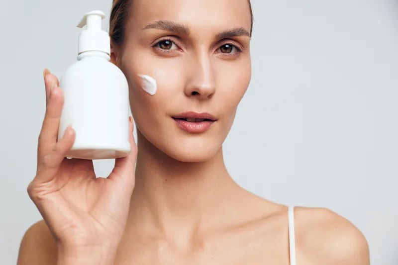 A young woman with clear skin holds a white lotion bottle near her face with a cream dot on her cheek. Concept for skincare, beauty routine, hydration, and facial care products.