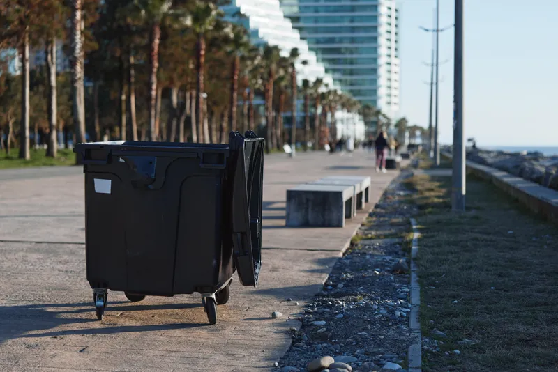 Distant pedestrians walk along a sunlit promenade while a large dumpster sits on the pavement, creating a stark contrast between daily life and urban clutter.