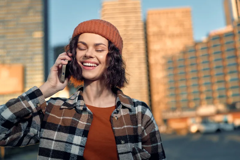 Woman on phone smile in urban sunset, authenticity and candid golden glow capture mindful lifestyle and emotional storytelling, cozy beanie and plaid jacket portrait of modern joy.