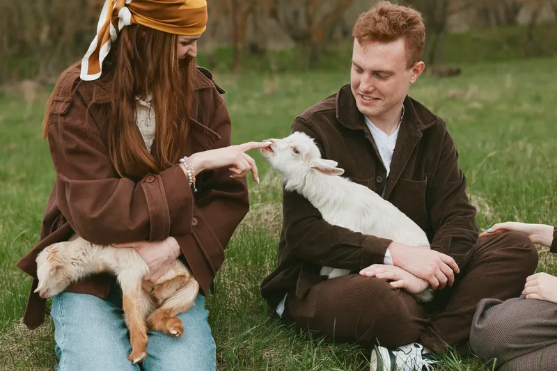 Goat kid with two people in a countryside field, gentle interaction and affectionate touch. Casual outdoor lifestyle scene highlighting companionship, animals, nature, and relaxation.