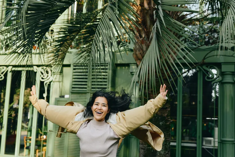Joyful woman with open arms in a sunny outdoor setting among palm trees, wearing a light sweater and jacket, conveying freedom, energy, and carefree celebration in a lush garden