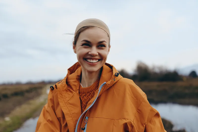 Woman lifestyle outdoor nature smile jacket river sky countryside travel portrait, smiling in orange jacket and cap near riverbank, countryside trail landscape, relaxed hike under soft daylight