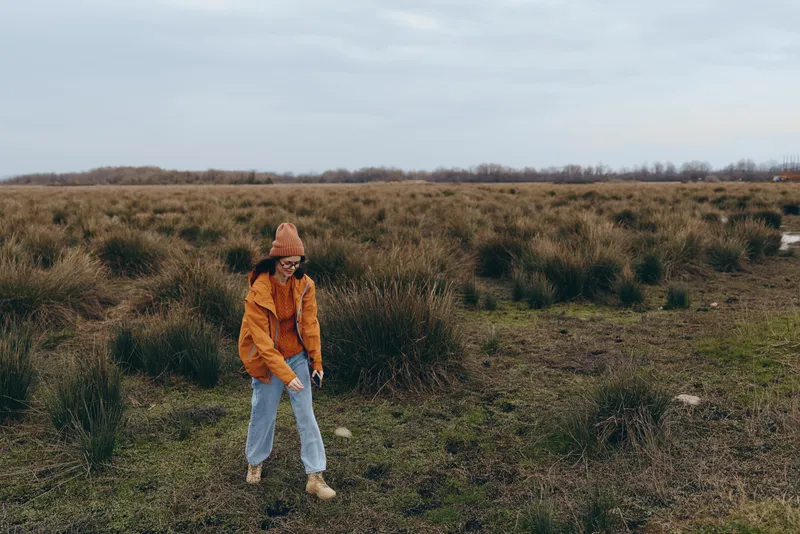 Woman lifestyle outdoors, field walking in tall grass, casual autumn look, orange sweater and knit hat, jeans, on dirt path, calm countryside mood, natural light