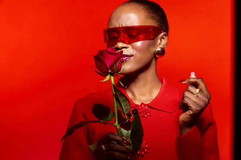 Elegant dark skinned African woman in red outfit and futuristic sunglasses enjoys the scent of a red rose. Concept of fashion, beauty, and passion with vibrant red background and accessories.