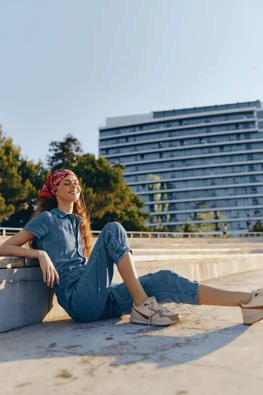 Lifestyle woman in modern boho-western denim street style relaxing outdoors under warm sunlight with a film color aesthetic and casual sneakers.