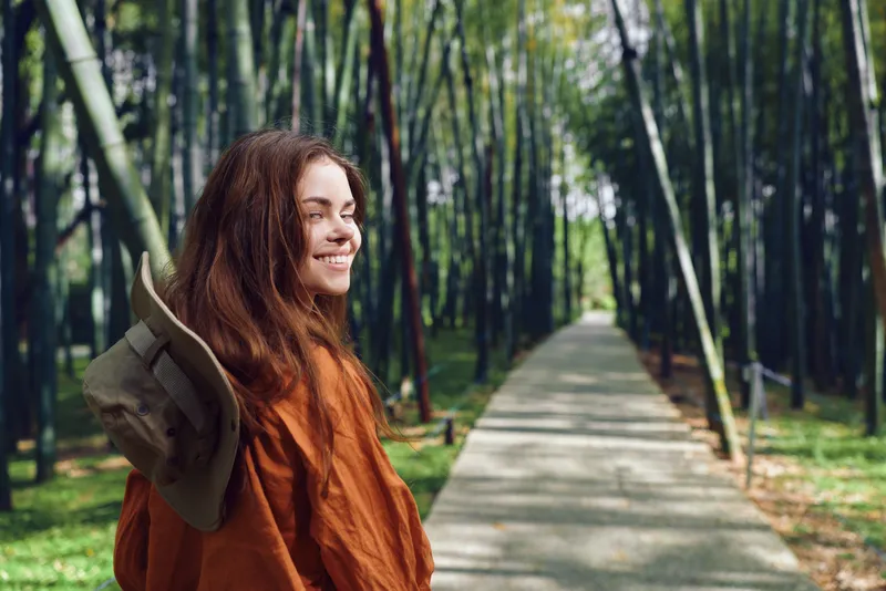 Woman smile in bamboo forest on a sunlit path wearing hat and orange jacket, portrait of a traveler enjoying travel and nature, peaceful green grove and outdoor leisure.
