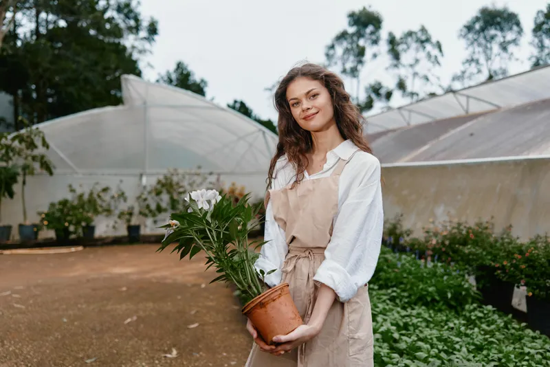Young woman with curly hair smiling while holding a potted plant in a greenhouse, wearing a beige apron and white blouse, showcasing a connection to nature