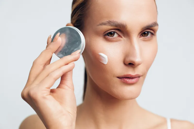 A young woman applies white cream on her cheek using her fingers while holding an open jar of skincare product. The background is plain and light, focusing on beauty care.