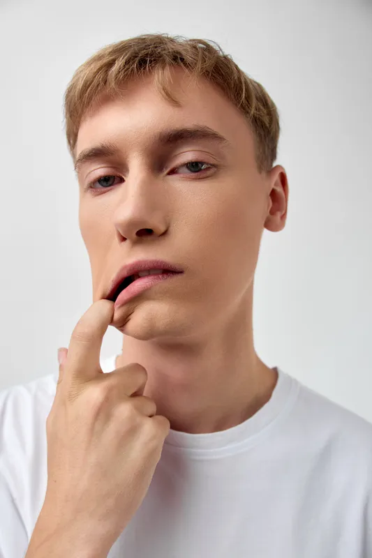 Close up of a young man with a thoughtful expression, finger near lips, subtle bite, wearing a white shirt in a minimal studio with soft lighting