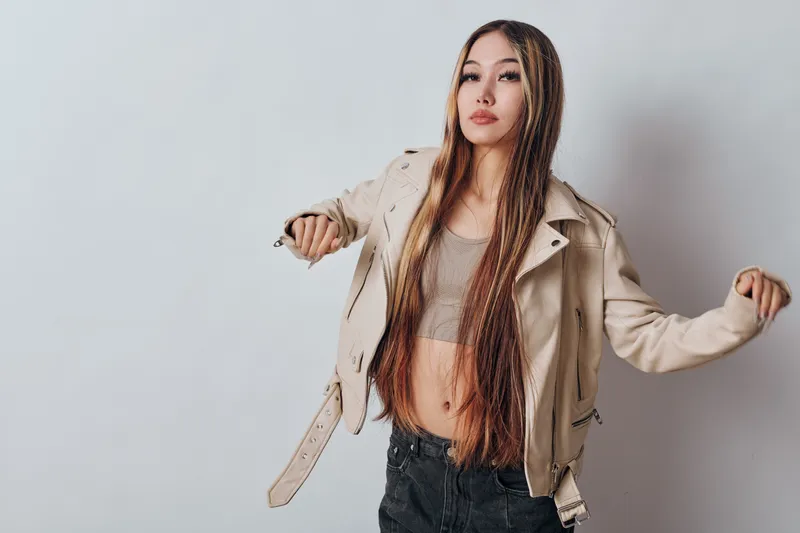 Young woman with long braids wearing beige jacket and crop top posing in studio with confident expression. Fashion model with casual outfit and trendy hairstyle on plain background.