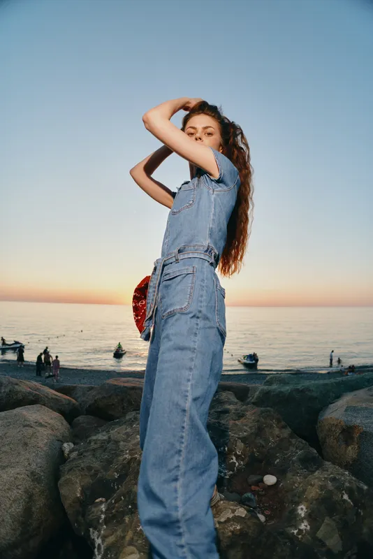 Lifestyle woman in modern boho-western denim street style stands confidently on rocky shore with film color tones during sunset by the sea.