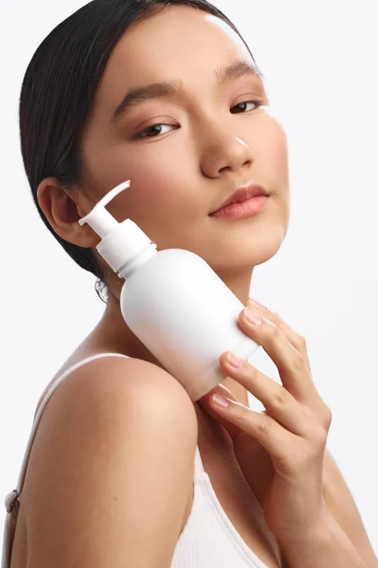 asian woman poses with a pump bottle in a soft studio setting, embodying skincare routine and self care. her serene expression and gentle light convey calm beauty and wellness.