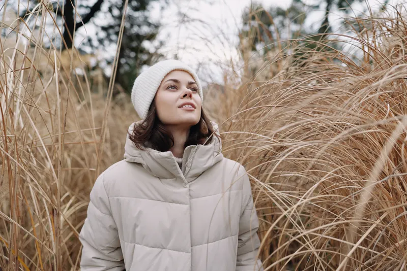Winter scene with a woman in a padded coat among tall dry grasses, gazing upward in a calm outdoor field and soft light adds calm mood.