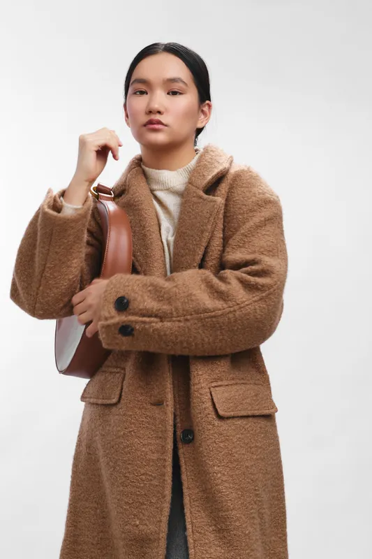 Asian woman in a camel wool coat stands in a clean studio, posing with a bag over her shoulder. She projects confidence and style, creating a calm, fashion forward atmosphere that feels contemporary
