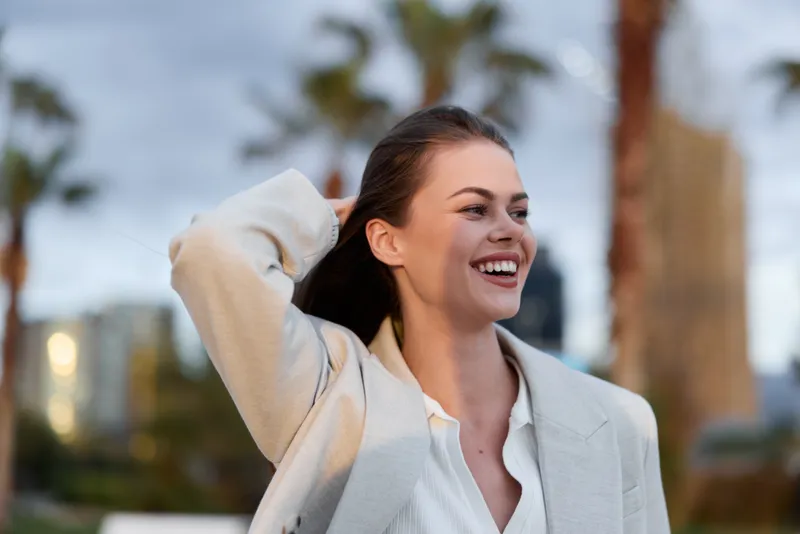 Cheerful woman smiling outdoors in light business jacket with blurred city and palm trees background in spring or summer